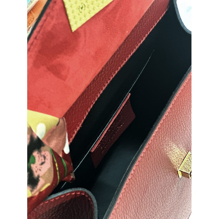 A red leather handbag with gold-tone metal details and a decorative scarf attached to the handle.