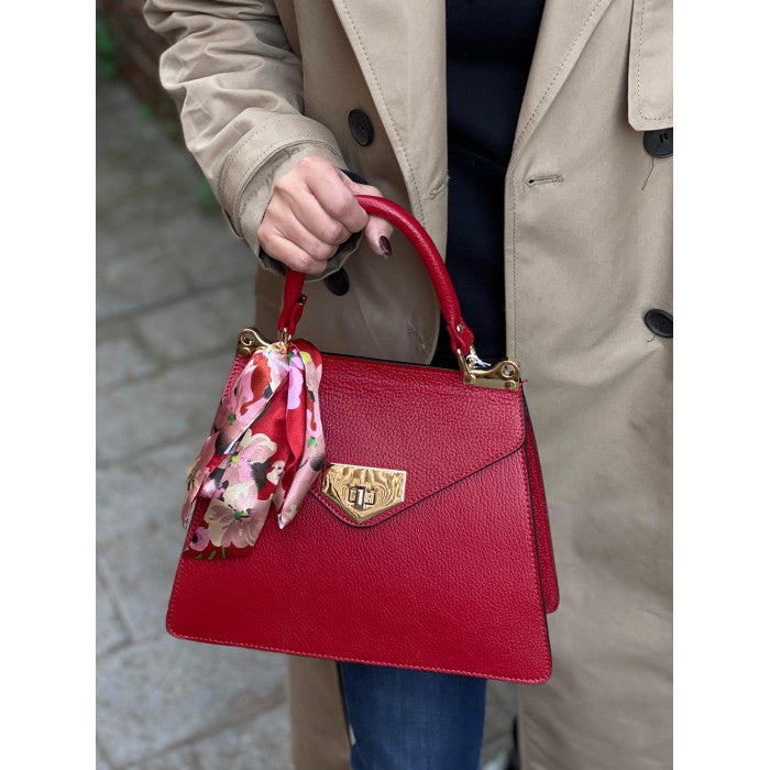 A person holding a red leather handbag with gold-tone metal details and a decorative scarf attached to the handle.